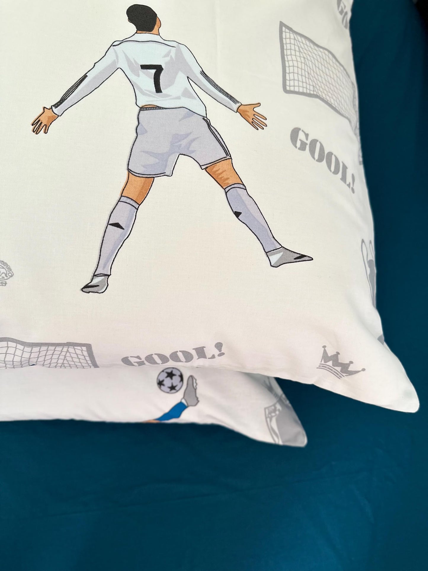 May include: White cotton pillowcases with a soccer theme. The design features a gray illustration of a soccer player with the number 7 on their jersey, goal nets, and the word 'GOOL!' printed in gray. A smaller illustration of a player kicking a soccer ball is also visible. A gray crown is printed on one pillowcase. The pillowcases are shown on a teal surface.