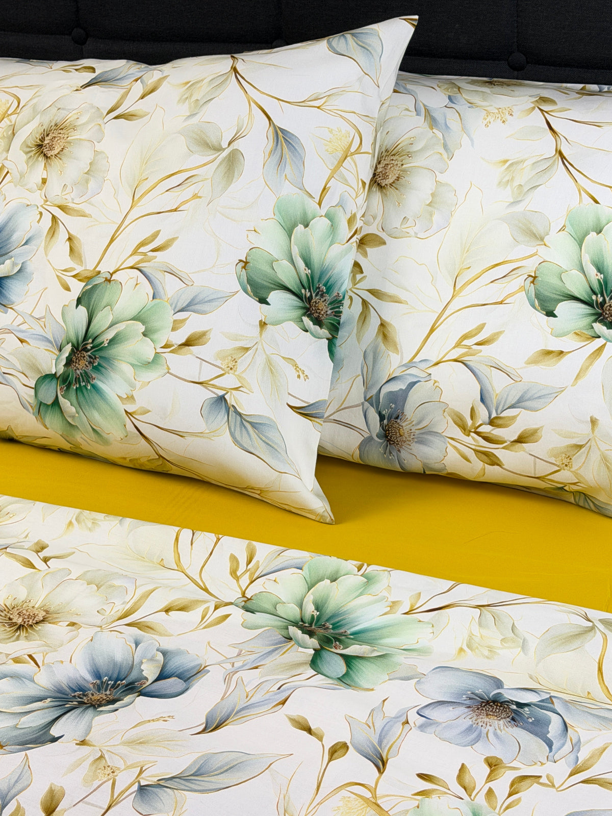 A close-up of a floral bedding set featuring two pillows and a matching duvet cover. The pattern showcases large flowers in shades of sage green, dusty blue, and soft yellow on a warm cream background, accented by delicate gold leaf-like veins.
