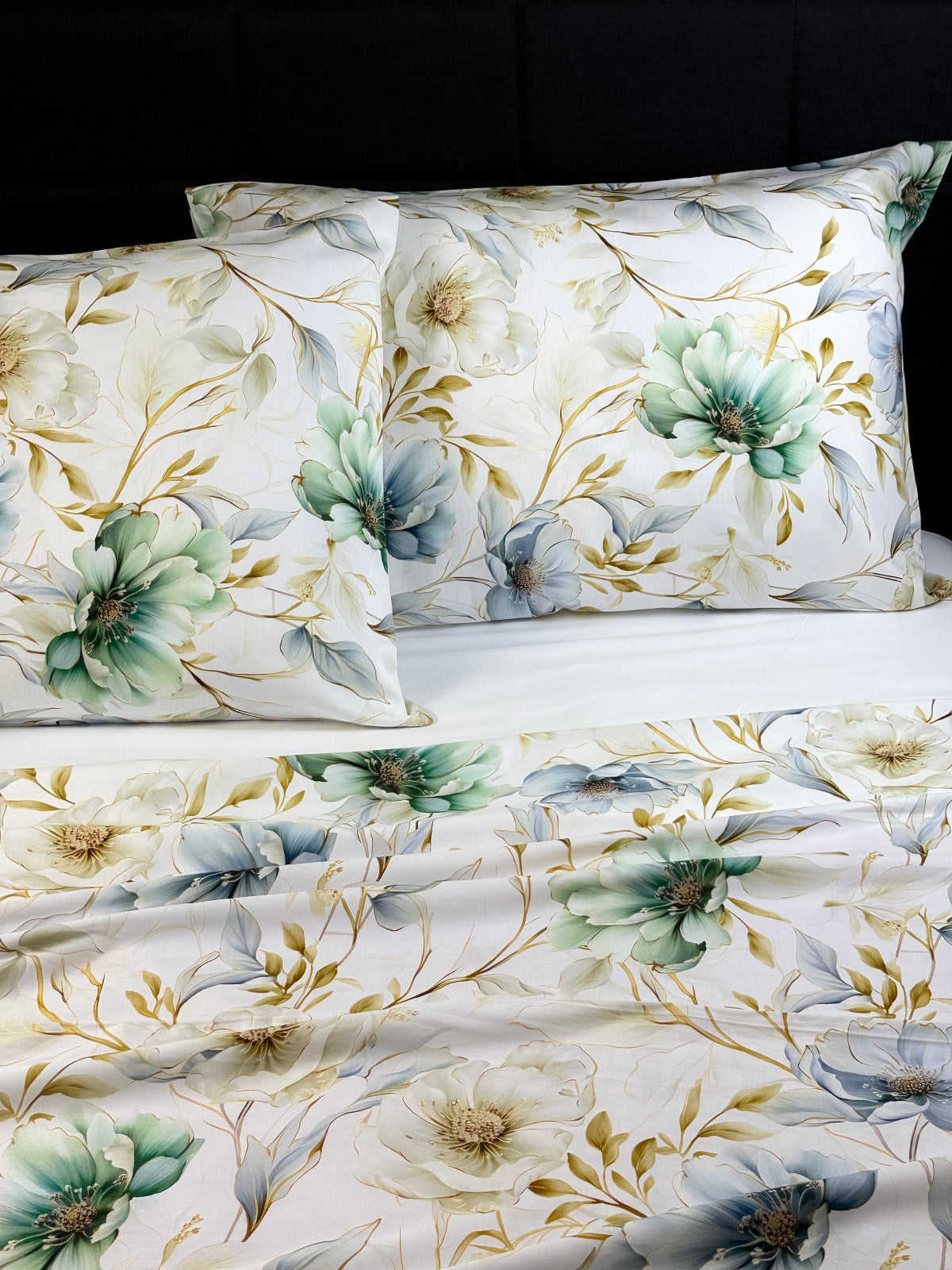A close-up of a bedding set featuring a floral print. The set includes two square pillows and a matching draped fabric panel, all in white with large blue-green and gray flowers, beige leaves, and gold-toned stems.