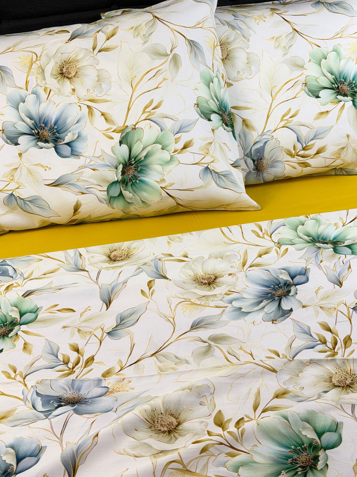 A close-up of a floral bedding set featuring cream-colored sheets and pillows with large blue, green, and white flowers, gold-toned leaves, and a bright yellow bedspread visible underneath.