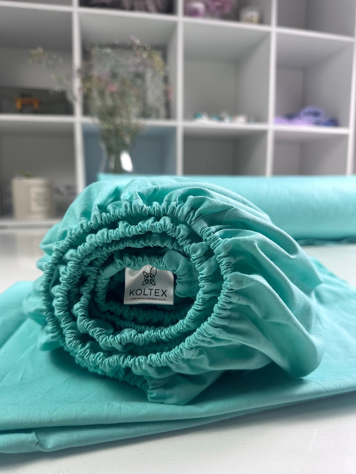Rolled teal cotton fitted sheet with elastic corners and Koltex brand label — soft, breathable bedding on white shelf